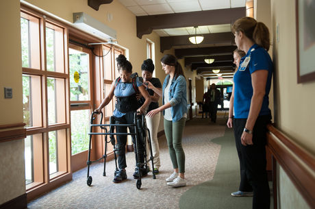 Black woman uses an assisted walking device while receiving physical therapy with the help of two other women in a hallway next to a window.